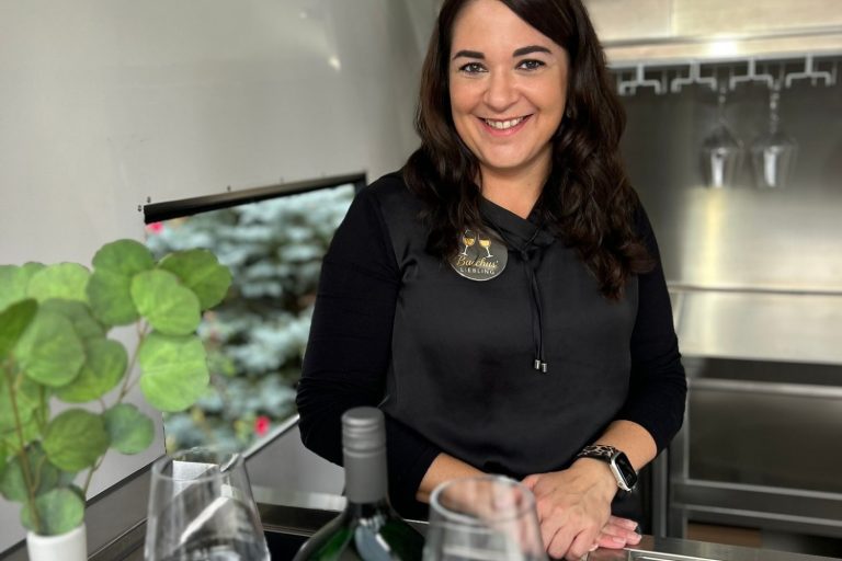 Frau in schwarzem Outfit lächelt neben einer Flasche Wein und Gläsern.